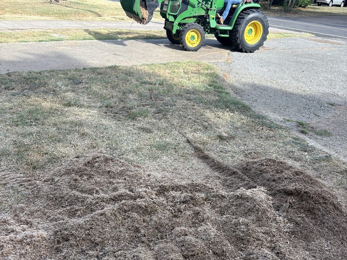A compact tractor on the drive with a loaded bucket raised, fresh grindings spread over the lawn in front.