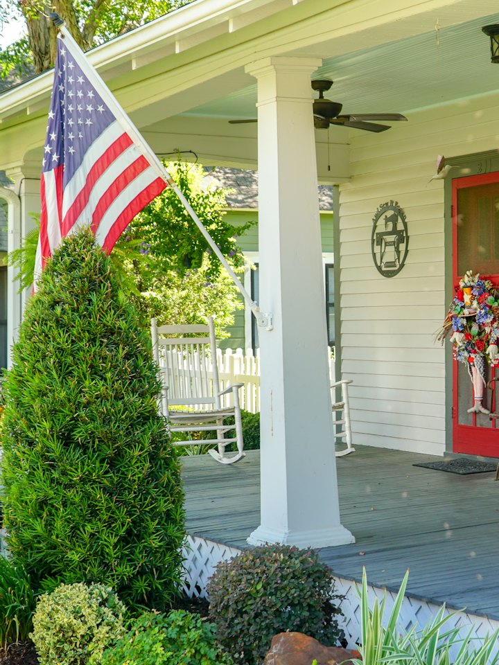 A Texas farmhouse porch flying the American flag, with white rocking chairs behind the column.